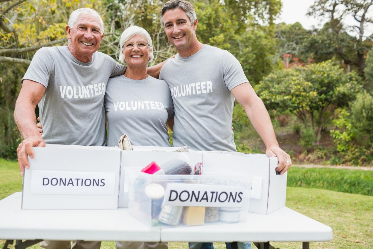 Three male and female senior volunteers stand outside behind table with donation boxes filled with