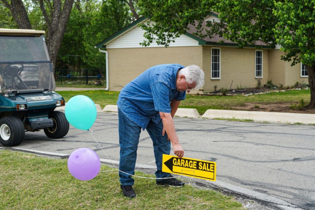 Senior man sets up "garage sale" sign with a purple and a blue balloon in his yard