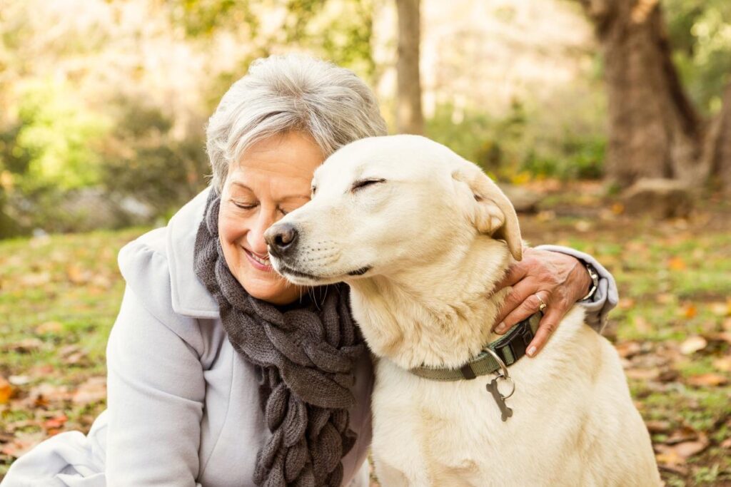 Mature woman smiles with her eyes closed as she hugs a blonde labrador retriever outside in the grass