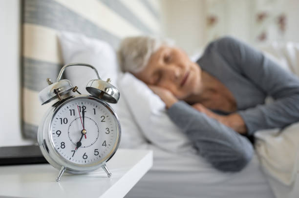 Senior in bed with a clock that reads 7 o'clock on her bedside table.