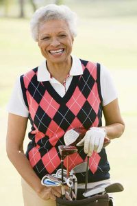 Mature black woman wearing golf attire smiles as she holds her bag clubs