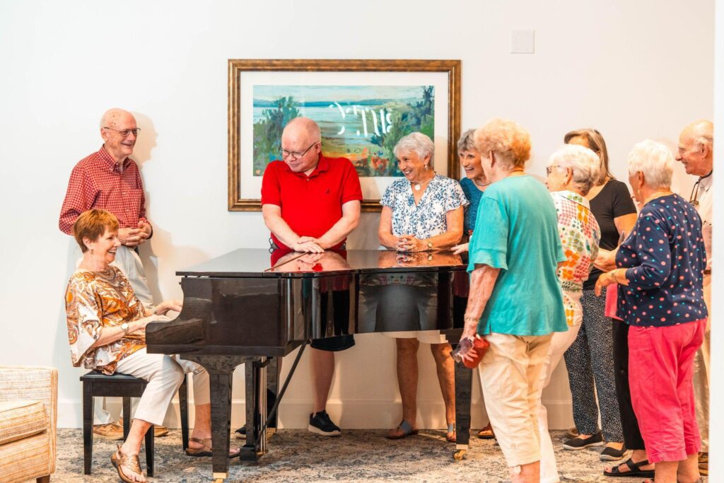 Senior residents gathering around a piano while an older woman plays at Porters Neck Village